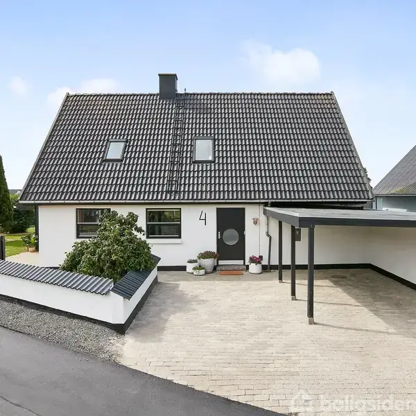 En hvid villa med skråt tag og carport, beliggende ved en vej. Nummer 4 ses ved indgangsdøren. Haven har buske og en indkørsel af lyse sten.