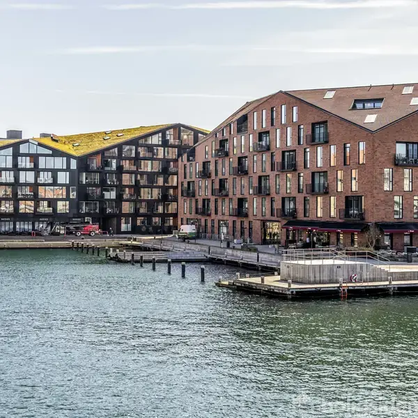 Moderne lejlighedskompleks ved en kanal med skrånende tage og store vinduer. Bygningerne har brune murstensfacader og grønne tage, mens vandet reflekterer lys fra en klar himmel.