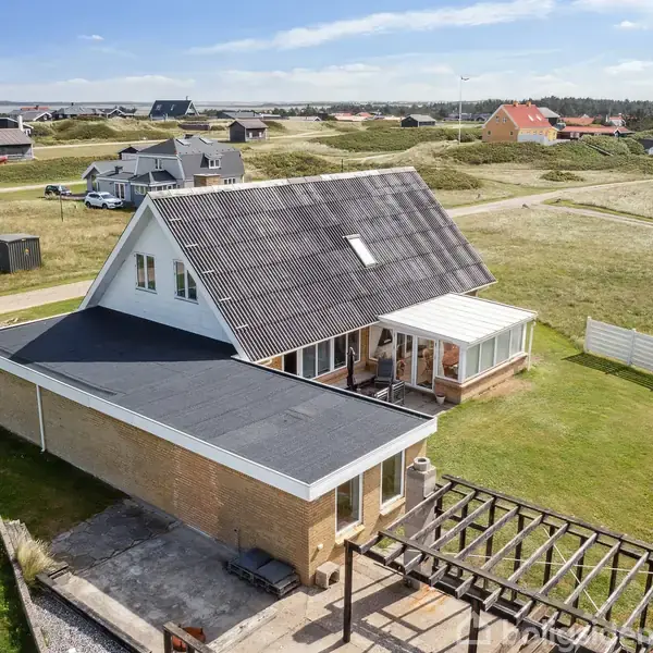 Et hus med skråt sort tag og tilhørende udestue ligger i et grønt, åbent område med flere andre huse i baggrunden. En pergola står foran huset.