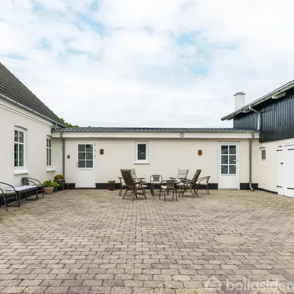 En flisebelagt gårdsplads med et sæt havemøbler i midten, omgivet af hvide bygninger med døre og vinduer, under en overskyet himmel.