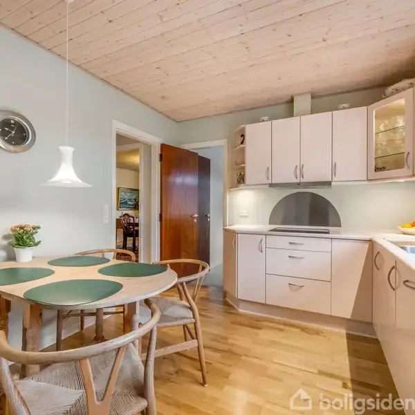Et lyst køkken med træbetrukket loft. Ovale bordplader på spisebordet med fire stole. Vægmonterede skabe og en moderne vask. Døren fører til en anden stue. Ingen tekst bortset fra "boligsiden".