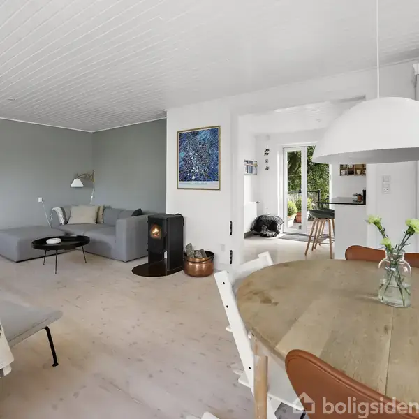 En lysegrå sofa står mod en væg med to billeder, ved siden af et rundt sofabord og en kamin. Rummet har et rundt spisebord med en vase i lyse omgivelser.