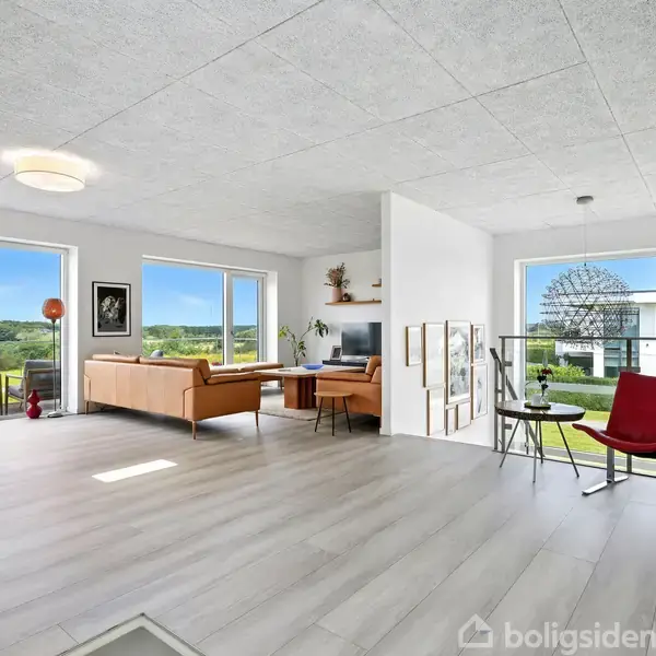 Stue med lyse trægulve, moderne møbler placeret mod store vinduer, der giver udsigt til en frodig have og et moderne hus. Indretningen er minimalistisk og lys.
