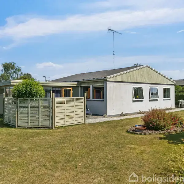 Et hvidt enfamiliehus står på en velplejet græsplæne med blomsterbede. Der er en terrasse foran huset og en solparasol i baggrunden. Himlen er klar og solrig.