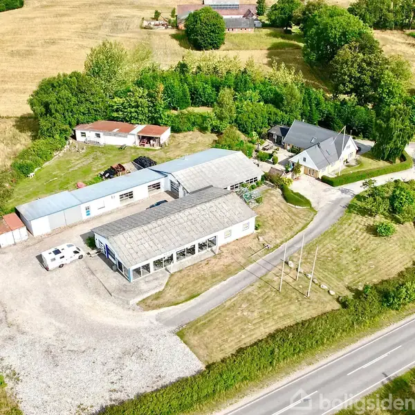 En række bygninger ligger i et landligt område med veje og grønt landskab omkring. Flere strukturer, herunder et drivhus og en bolig, omgiver et åbent grusareal.