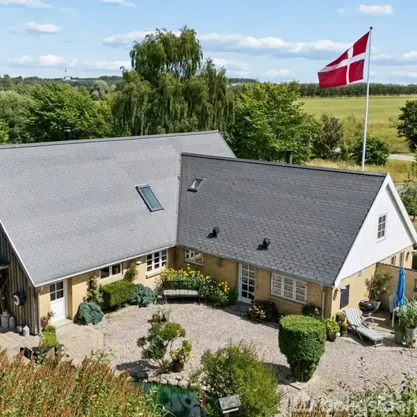En gul murstensbygning med grå tage ligger i en velplejet have. En stor dansk flagstang med flag står i nærheden, omgivet af grønne marker og træer under en blå himmel.