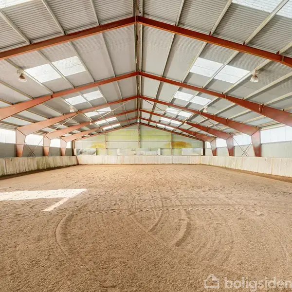 Ridehal med sandbund ligger stille under et stort metaltag med røde bjælker; omgivet af hvide barrierer og naturligt lysindfald fra ovenlysvinduer.