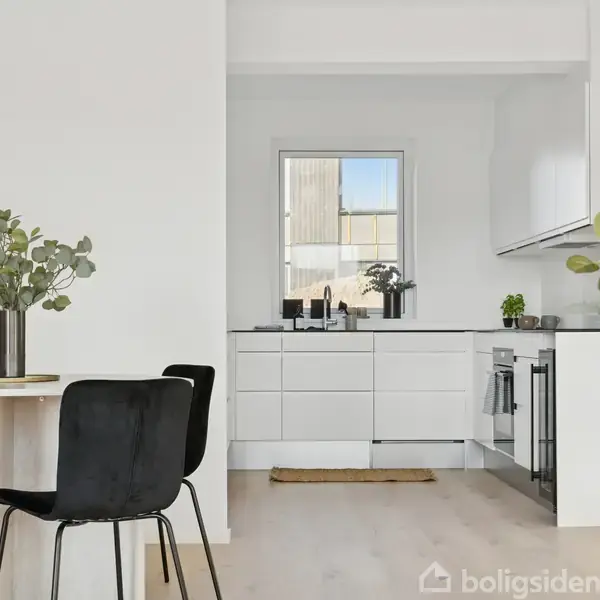 Køkkenbord med sorte stole står ved siden af et hvidt, moderne køkken med vask under et vindue. Grønne planter pynter både bordet og køkkenet.