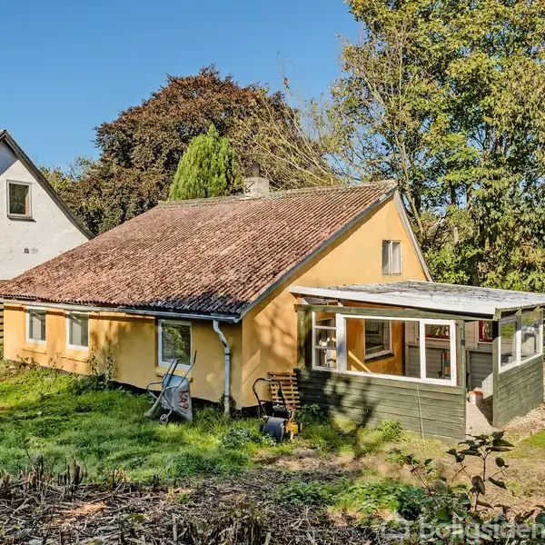 Et lille, gult hus med skråt tag står stille i en solrig have. Omkring huset er der grønne træer og buske. Til venstre ses et nærliggende hvidt hus.