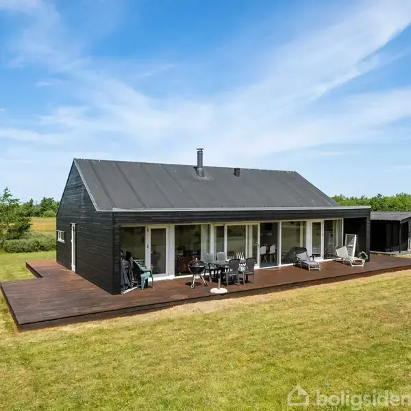 Et sort enfamiliehus med store vinduer står på en træterrasse. Havemøbler er placeret foran huset. Omgivet af en grøn græsplæne og træer under en klar blå himmel.