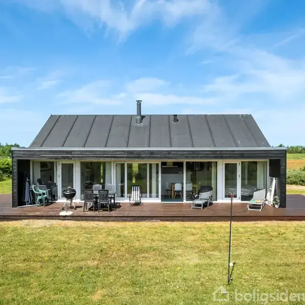 Et moderne sommerhus med sort tag, store glasfacader og en træterrasse. Der står havemøbler på terrassen. Huset er omgivet af en grøn græsplæne og skovklædt baggrund.