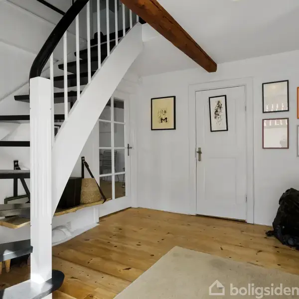 Trappeopgang med sorte trin og hvidt gelænder fører opad, omgivet af hvidmalede vægge med billedrammer. Trægulv og en lukket dør. Atmosfæren er hjemlig og moderne.