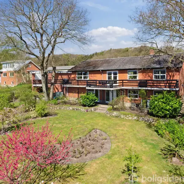 Rødt hus med sort tag ses fra haven, omkranset af store træer og blomstrende buske. Foran huset er der en velholdt græsplæne, og i baggrunden ses en skov.