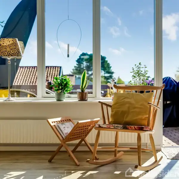 En gyngestol med pude står ved et stort vindue, der giver udsigt til en solrig terrasse med planter. En magasinholder med magasiner står ved siden af stolen.