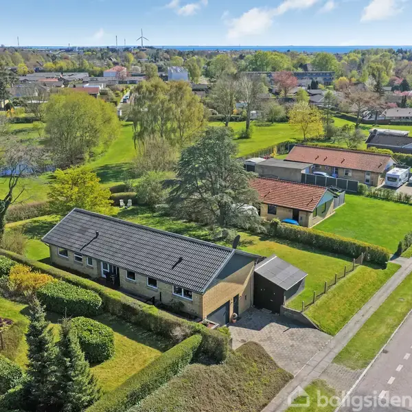 En enkeltfamilieshus med mørkt tag står blandt velplejede haver og grønne træer. Der er en flisebelagt indkørsel, og i baggrunden ses flere huse og åbne græsarealer.