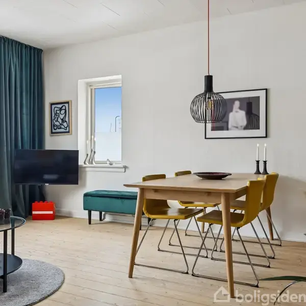 Stue med spisebord og fire gule stole står ved en hvid væg med indrammet kunst. Et rundt sofabord står på tæppe nær grønlige gardiner. En lampe hænger over bordet.