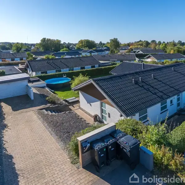Et parcelhus med sort tag er omgivet af en hæk i et villakvarter. Foran huset står affaldscontainere ved en flisebelagt indkørsel. Baggrunden indeholder flere huse og træer.