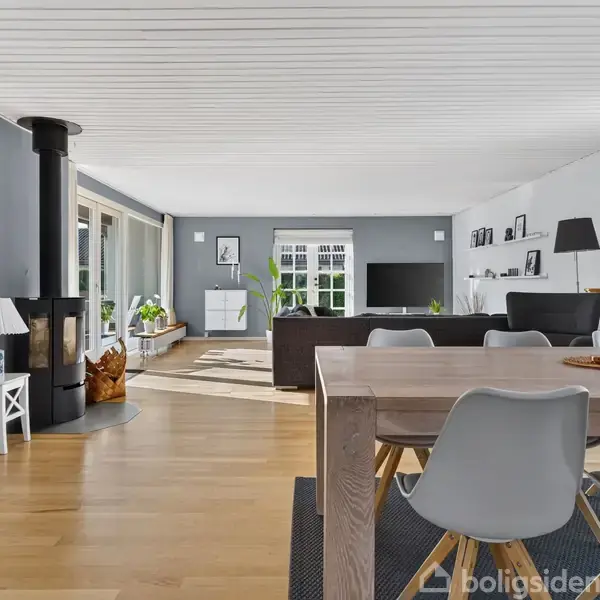 Et lyst stueområde har moderne indretning med spisebord, stole og sofa. En brændeovn står til venstre. Store vinduer giver udsigt til haven og naturligt lysindfald.