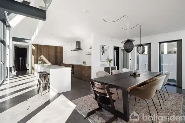 Moderne køkken-alrum med træbord og stole omgivet af store vinduer. Køkkenø med barstole står ved siden af mørkebrune trægarderober. Lysindfald kommer fra ovenlys og vinduer.