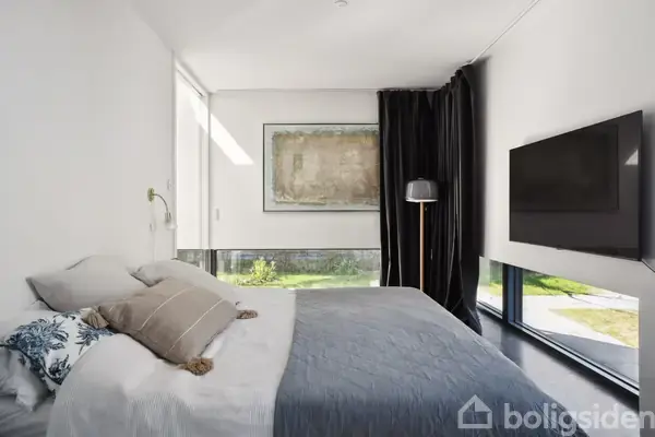 En seng med flere puder står i et lyst værelse. Ved siden af hænger et maleri på væggen. Til højre ses en stor fladskærm, og vinduer giver udsigt til have.