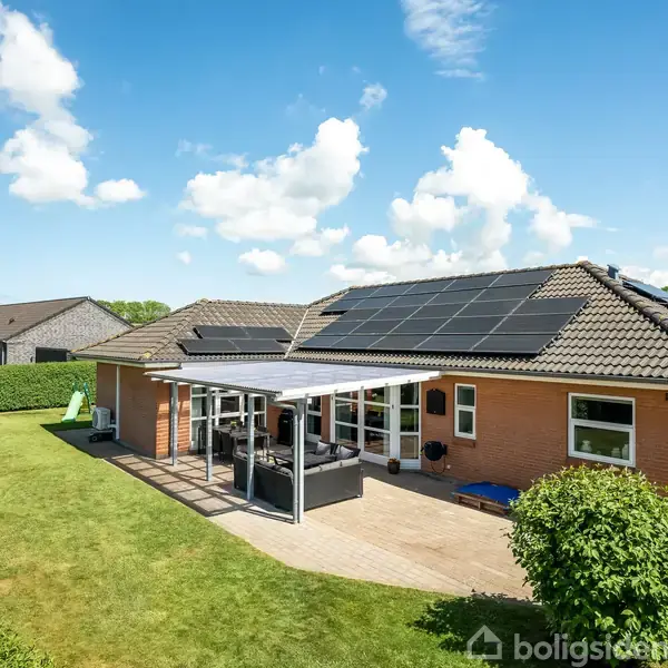 En murstensvilla med solpaneler på taget. En overdækket terrasse med havemøbler er foran huset. Græsplæne med en gynge til venstre. Baggrunden har en klar blå himmel.