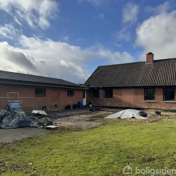 Et murstenshus med mørkt tag står på en græsplæne. Til venstre ses en carport og diverse objekter. Himlen er delvist skyet.