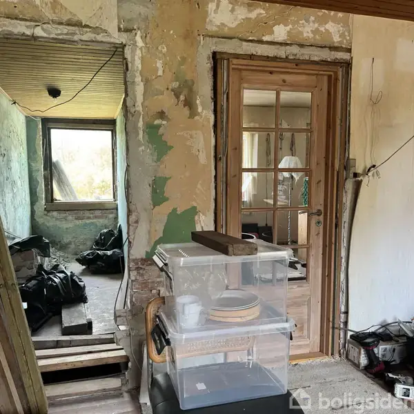 En delvist renoveret indendørs scene viser to rum forbundet af en dør. Et rum har ujævne vægge og et vindue. Det andet rum har en glasdør og gennemsigtige opbevaringskasser med tallerkener.