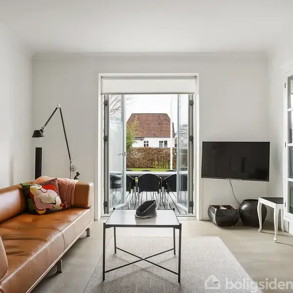 En moderne stue med en brun lædersofa står ved en glasskydedør. Et lille bord og en fladskærm på væggen. Udenfor ses et bord og stole på terrassen.