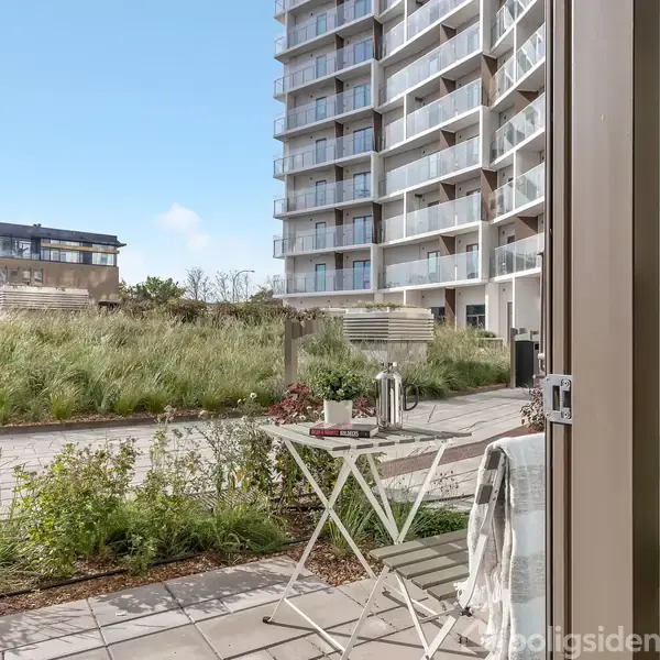 Et lille bord med en lanterne og kop står på en terrasse. I baggrunden ses en moderne beboelsesejendom ved en sti omgivet af grønne buske.