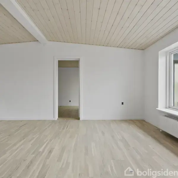 Værelse med lys trægulv og hvidt loft, rummelig og tom. To døråbninger i væggen, store vinduer til højre med udsigt til natur. Ingen tekst til stede.