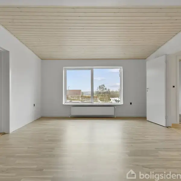 Et lyst rum med trægulv og hvidmalede vægge. Et stort vindue viser udsigt til træer og bygninger udenfor. Døre fører til andre rum. Ingen tekst ses på billedet.