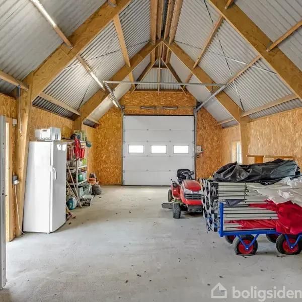 Garage med åben port. Indeni er en græsslåmaskine, en vogn med sikkert dækket udstyr og en hvidt køleskab til venstre. Rummet er lyst, med høj loftshøjde og trægitter.