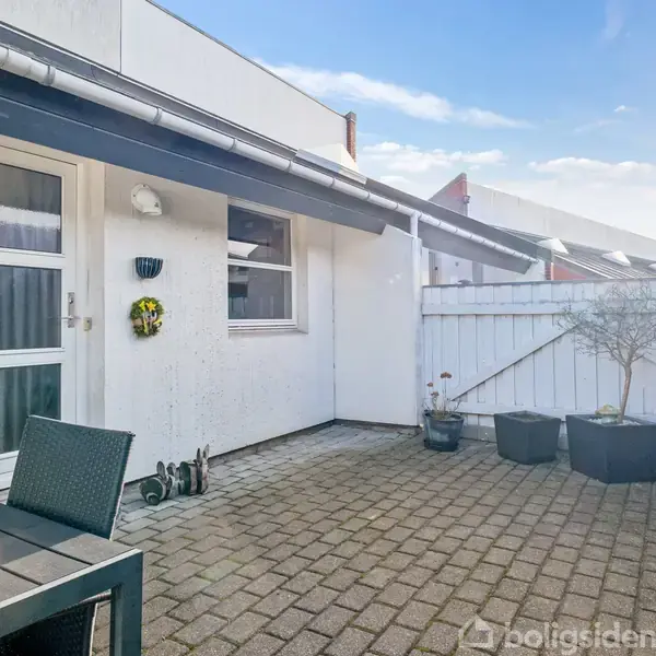 Terrassemøbler står på en flisebelagt gårdhave. Et hvidt hus med vindue og dør danner baggrunden. Et hvidt stakit omkranser området, og der er potteplanter i hjørnet.