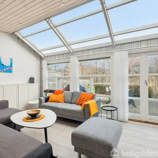 Grå sofa og bord med appelsinskuen dekorationer står i en stue med glasloft og store vinduer, der giver udsigt til en træterrasse og have. Klar himmel udenfor.