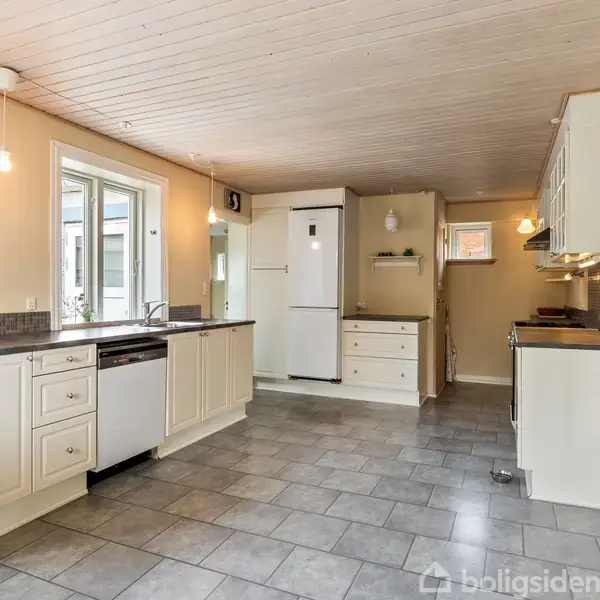Et moderne køkken med hvide skabe og grå flisegulv. Køleskab og opvaskemaskine står langs væggen. Store vinduer giver naturligt lys. En dør fører til en anden del af huset.
