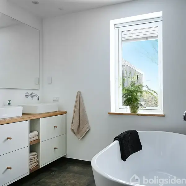 Et lyst badeværelse med et badekar ved siden af et vindue, en håndvask på en træbordplade og skabe nedenunder. En håndklædeholder og planter skaber en afslappet atmosfære.