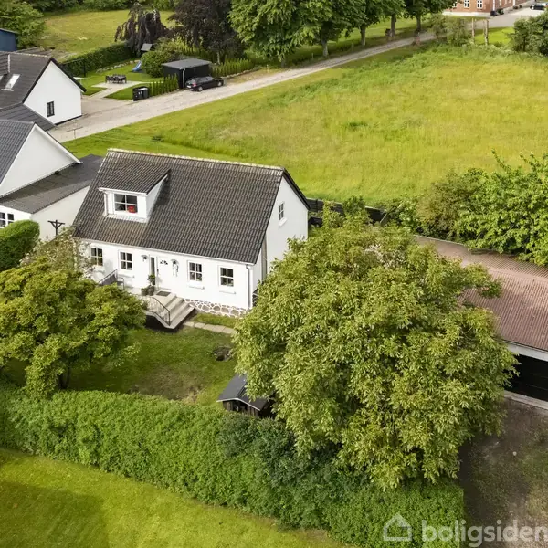 Hvidt hus med skråtag står ved siden af en lade i en have omgivet af tæt vegetation. Husene ligger i et grønt, landområde med stier og andre boliger i nærheden.