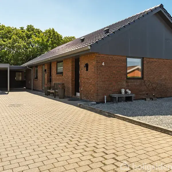 En enkeltstående rødstensvilla med grå tag, beliggende i solrigt vejr. Foran huset er en bred flisebelagt indkørsel. Tallet "4" er tydeligt på huset. Spredte buske langs siden.