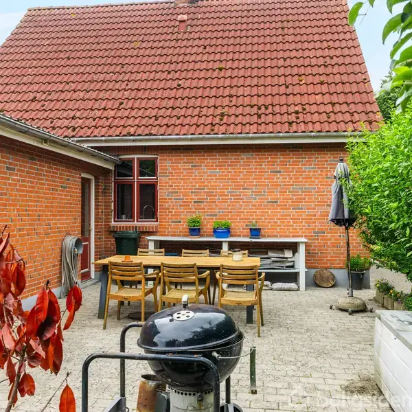 Grill står klar på en gårdsplads. Et bord med stole er placeret foran en rød murstensbygning med et rødt tegltag. Omgivet af grønne planter og buske.