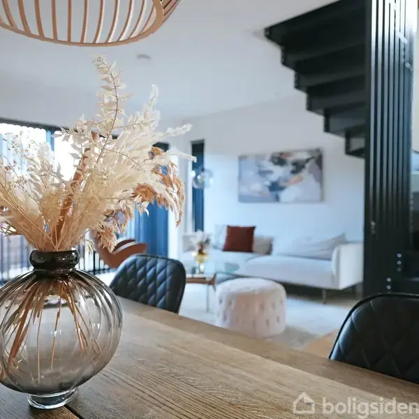 En vase med tørrede blomster står på et spisebord i en stue. I baggrunden ses en sofa, gulvpuder og en moderne trappe.
