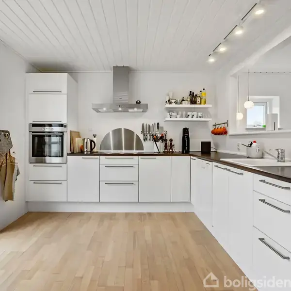 Et moderne hvidt køkken med træbordplader. Der er en indbygget ovn, emhætte, og åbne hylder med køkkenredskaber. Store vinduer giver naturligt lys. Vandhane og vask ses i højre side.