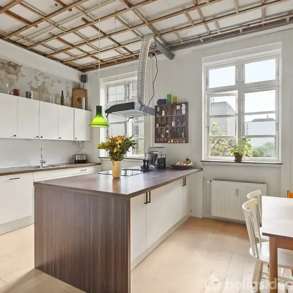 Køkken med hvidt skab og ovn, hvor der står en vase med blomster på mørkt køkkenbord. Grønne lamper hænger fra loftet. Lyst rum med stort vindue.