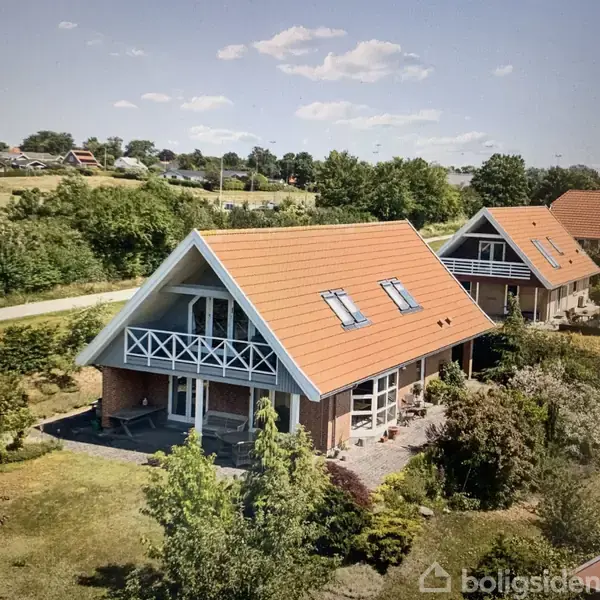 Hus med rødt tegltag og store vinduer står i en velplejet have. Balkonen har hvidt gelænder. Baggrunden viser et grønt, landligt område med flere huse.