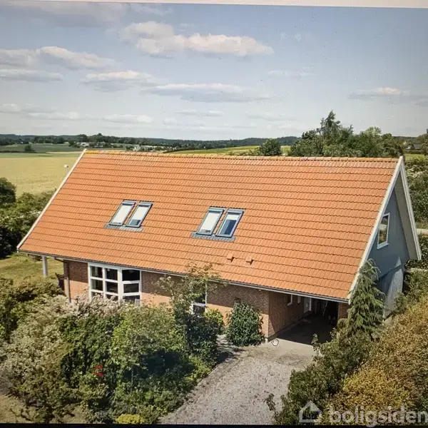Et rødt tegltagshus med to ovenlysvinduer står omgivet af frodig vegetation og en grusvej i en landlig setting med marker og skov i baggrunden.