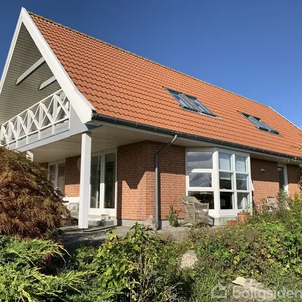 Et rødt teglhus med skråt tag og flere ovenlysvinduer står i en frodig have med buske og træer. Huset har en altan og en terrasse med stole.