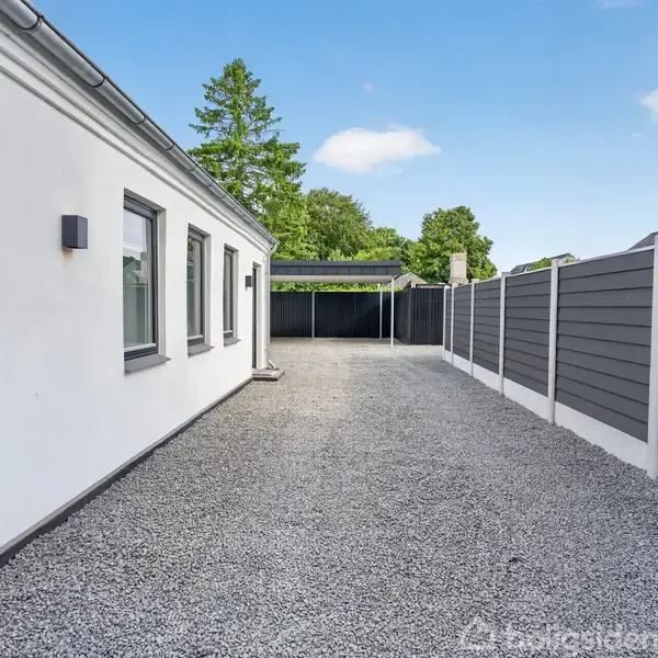 Hvid bygning med flade tag og vinduer langs siden støder op til en grusbelagt indkørsel. Grå plankeværk på den anden side af indkørslen. Træer og carport i baggrunden.