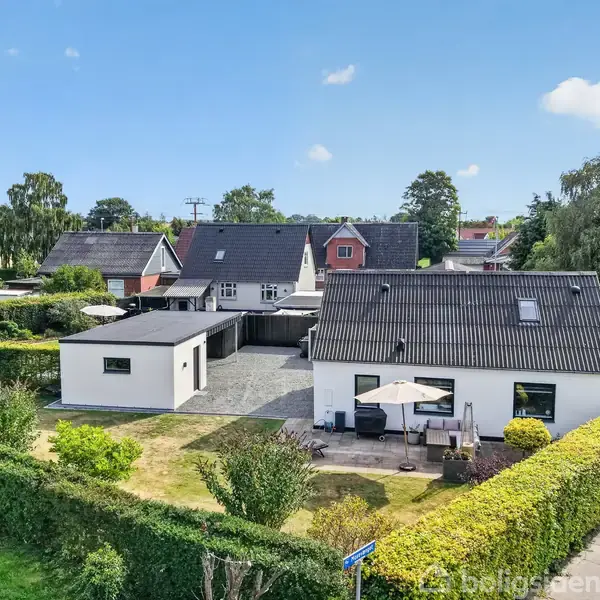 Et hvidt hus med sort tag står i en velplejet have med terrasse og parasol. Omgivet af andre huse og træer i et villakvarter.