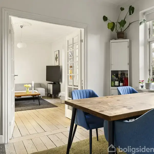 En spisebord fyldt med blå stole står i en spisestue ved siden af et stort vindue med planter. En åbning fører til en stue med sofa og tv i lyse omgivelser.