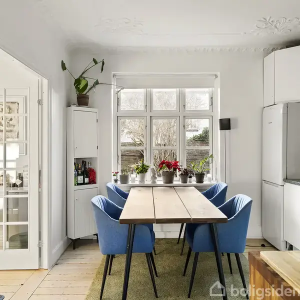 Et spisebord med seks blå stole står i et lyst køkken. Bordet er placeret foran et stort vindue, mens et hvidt skab og en grøn væg ses i baggrunden.