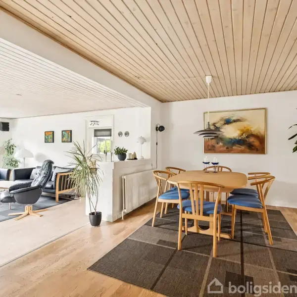 Spisestue med bord og seks stole placeret på tæppe. Bagved er en sofa og lænestol i en lys stue. Vægdekorationer og planter skaber en hyggelig atmosfære.